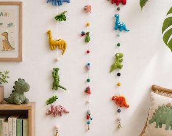 May include: Three vertical hanging garlands featuring felt dinosaur figures in blue, yellow, green, red, and pink, interspersed with small felt balls in various colors. The garlands are suspended from a white wall and include small metal bells at the bottom.
