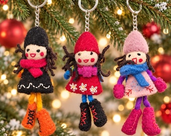 May include: Three felt doll keychains hanging from a tree. The left doll wears a black hat and coat with a red scarf and orange boots. The center doll has a red hat and coat with a purple scarf and black boots. The right doll has a pink hat and coat with a blue scarf and pink boots.