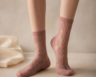 May include: A pair of sheer, dusty rose socks with a floral lace pattern. The socks have a ribbed cuff and are worn on a person's legs. The background is a neutral color.