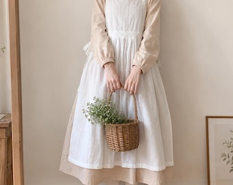 May include: A beige long-sleeved dress with a rounded collar paired with a white sleeveless apron featuring a gathered waist. The outfit is complemented by a small, round, woven brown wicker basket filled with green foliage.