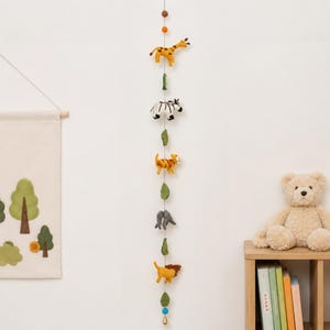 Puede incluir: Una guirnalda vertical colgante con figuras de fieltro de una jirafa, cebra, tigre, elefante y le&oacute;n, intercaladas con hojas de fieltro verde y peque&ntilde;as bolas, suspendidas en un hilo fino con una peque&ntilde;a campana en el extremo inferior.