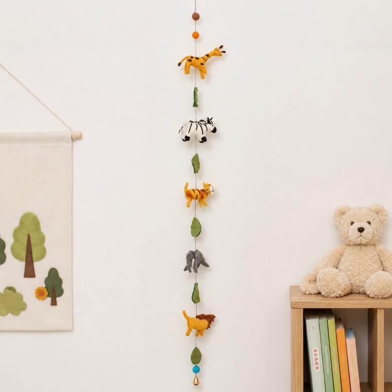 Puede incluir: Una guirnalda vertical colgante con figuras de fieltro de una jirafa, cebra, tigre, elefante y le&oacute;n, intercaladas con hojas de fieltro verde y peque&ntilde;as bolas, suspendidas en un hilo fino con una peque&ntilde;a campana en el extremo inferior.