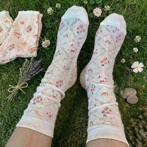 May include: A pair of white socks with a floral pattern of pink roses and green leaves. The socks have a diamond-shaped openwork design and are laying on green grass.