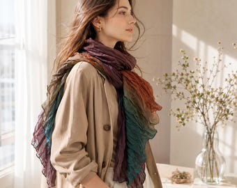 May include: A long, lightweight scarf featuring a gradient of deep purple, burnt orange, and dark teal colors. The fabric has a crinkled, textured finish and is draped loosely around the neck, worn over a tan blazer and light-colored trousers.