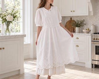 May include: A white midi-length dress featuring short puff sleeves, a round neckline, and a fitted bodice. The skirt is layered with a sheer overlay adorned with intricate white floral embroidery and a scalloped lace hemline detail.