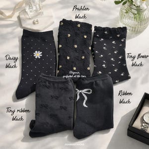 May include: Five pairs of black socks with various patterns, including daisies, tiny flowers, bows, and textured designs. The socks are labeled "Daisy black", "Prahlen black", "Tiny flower black", "Tiny ribbon black", and "Ribbon black". The text "Elegance, perfected at the toes" is also visible.