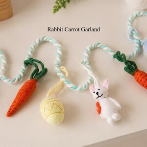 Puede incluir: Una guirnalda Rabbit Carrot Garland con una cuerda trenzada en blanco y azul claro, decorada con zanahorias de fieltro naranja con hojas verdes, un huevo amarillo p&aacute;lido con patr&oacute;n en zigzag y un peque&ntilde;o conejo blanco con rasgos negros.