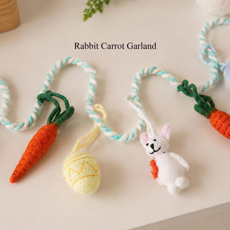 Puede incluir: Una guirnalda Rabbit Carrot Garland con una cuerda trenzada en blanco y azul claro, decorada con zanahorias de fieltro naranja con hojas verdes, un huevo amarillo p&aacute;lido con patr&oacute;n en zigzag y un peque&ntilde;o conejo blanco con rasgos negros.