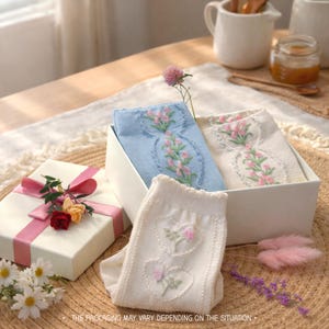 May include: A white gift box with a pink ribbon and floral decorations is next to a white box containing light blue and off-white embroidered socks. The socks have floral designs. Daisies and other decorative elements complete the scene.