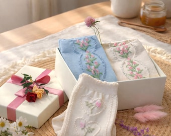 May include: A white gift box with a pink ribbon and floral accents sits next to a white box containing light blue and off-white embroidered socks. The socks feature floral designs. Daisies and other decorative elements complete the scene.