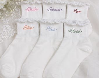May include: A set of three pairs of white ankle socks with ruffled cuffs. Each pair features embroidered text in different colors: pink 'Bride', purple 'Forever', red 'Love', orange 'Bliss', light blue 'Vow', and green 'Cherish'.
