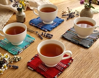 May include: Four white teacups filled with tea rest on colorful, square coasters. The coasters are blue, green, gray, and red, with a woven texture. Small vases of flowers and sprigs of lavender decorate the wooden table. The text at the bottom says: It is a handmade product, and the color may vary slightly depending on the store of production.