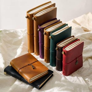 Puede incluir: Una colección de diarios de cuero en varios colores, incluyendo marrón, morado, verde y rojo. Cada diario tiene un cierre de correa y un dije decorativo. Los diarios están apilados y dispuestos sobre una superficie blanca.