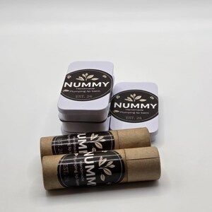 May include: A collection of white rectangular tins and brown lip balm tubes. The tins feature a black oval label with the word "NUMMY" and the text "Handmade plumping lip balm EST. 24". The lip balm tubes have a similar label with the text "UM NUMMY".