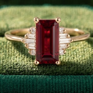 May include: A gold ring with a rectangular, deep red gemstone as the focal point. The ring is accented with smaller, baguette-cut white gemstones and small round diamonds. The ring is set against a green velvet background.