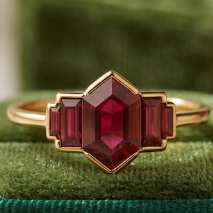 May include: A gold ring with a central, faceted, deep red gemstone in a geometric design. Two smaller rectangular red stones flank the main stone, all set in a gold band. The ring is presented on a green velvet surface.