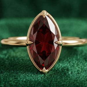 May include: A gold ring featuring a large, marquise-cut deep red gemstone set in a four-prong setting. The band is smooth and polished, showcasing a classic design that highlights the elongated shape and faceted surface of the central stone.