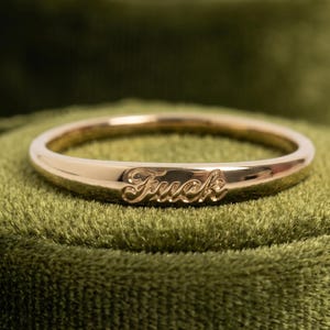 Puede incluir: Un anillo dorado con la palabra "Fuck" grabada. El anillo se muestra sobre un fondo de terciopelo verde, resaltando su acabado pulido y diseño. Diámetro aproximado de 2 cm.