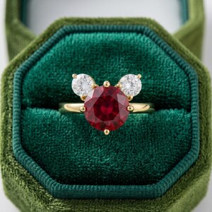 May include: A gold ring featuring a large, round, deep red gemstone flanked by two smaller, round, clear gemstones. The ring is set against a dark green velvet background, suggesting a jewelry box.