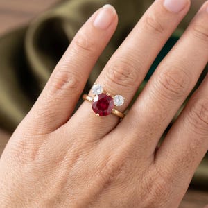 May include: A gold ring featuring a large, round, deep red gemstone as the centerpiece, flanked by two smaller, clear, round gemstones. The ring is set against a neutral background, highlighting the jewelry's design and color.