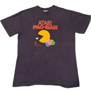 Puede incluir: Una camiseta negra de manga corta con un estampado gráfico del logotipo de Atari Pac-Man en texto rojo y naranja. Debajo del texto hay un personaje Pac-Man amarillo, tres fantasmas coloridos en azul, rosa y verde, y un pequeño texto de Atari.