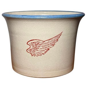 May include: A cream-colored ceramic planter with a wide, flared rim featuring a blue glazed edge. The front of the vessel displays a detailed, single red graphic of a stylized bird wing with intricate feather line work across the center.