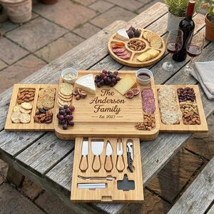 May include: A bamboo cheese board with the text "The Anderson Family Est. 2023" engraved on it. The board is open, revealing compartments filled with cheese, crackers, nuts, and fruit. A drawer holds cheese knives and a corkscrew.