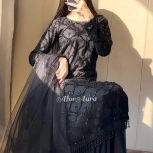 May include: Black formal dress with detailed beadwork and a flowing skirt. The long-sleeved top has a diamond pattern of beads. The dress includes a sheer, flowing shawl. The text "Afor Aura" is visible on the dress.