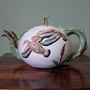 May include: A white ceramic teapot featuring a raised relief design of two flying ducks with brown and blue wings. The spout and handle are sculpted to resemble green cattail reeds with brown stalks, and the lid has a small leaf-shaped handle.
