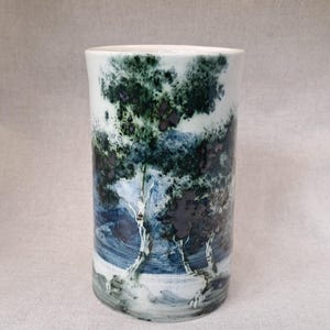 May include: A cylindrical ceramic vase featuring a hand-painted landscape scene with dark green and blue trees against a white background. The artwork depicts a forest setting with visible brushstrokes, creating a textured appearance on the surface.