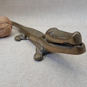 May include: A brass nutcracker shaped like an alligator with a textured body and a hinged jaw mechanism. The alligator is positioned with its mouth slightly open, and a single brown, textured walnut is placed near the tail of the metal figure.