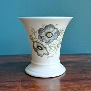 May include: A white ceramic vase with a flared rim and a wide base, featuring a detailed botanical illustration of two blue and grey anemone flowers with green stems and leaves centred on the front of the smooth, glossy surface.