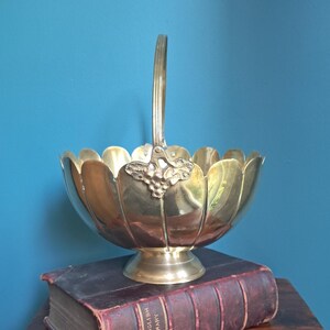 May include: A polished brass basket with a scalloped rim and a single upright handle. The base of the handle features a decorative grape and leaf motif. The basket rests on a pedestal base and is positioned atop a thick, worn, dark red book.