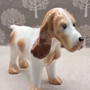 May include: A ceramic dog figurine, predominantly white with brown patches. The dog features long, drooping ears and a black nose. The figurine is posed on all fours, with its head slightly angled downwards.