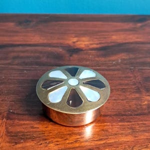 以下が含まれることがあります： 蓋に花柄のインレイが施された円形の真鍮製容器。白いマザーオブパールとダークブラウンの貝殻の交互の模様が、中央の白い円形のインレイの周りに配置されており、金属の表面に装飾的なデザインを作り出しています。