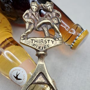 May include: A brass bottle opener with the words "THIRSTY PIXIES" and a design of three pixies. The opener is in front of two bottles of beer. The opener is a unique barware item.