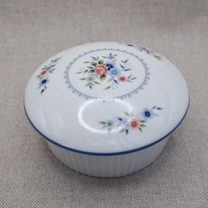 May include: A round white porcelain trinket box with a fluted base and a blue rim. The lid features a central floral bouquet of pink and blue flowers enclosed in a delicate grey circular border, with additional floral sprigs scattered around.
