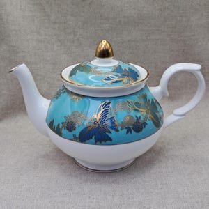 Könnte beinhalten: Eine weiße Teekanne aus Porzellan mit einem leuchtend türkisfarbenen Band um die Mitte und den Deckel. Der türkisfarbene Bereich ist mit goldenen und dunkelblauen Schmetterlings- und Blumenmotiven verziert. Der Deckel hat einen goldenen Knauf.