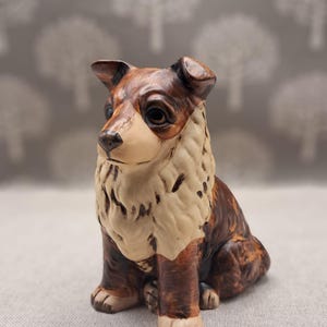 Può includere: Statuetta di cane in ceramica in posizione seduta. Il cane ha pelo marrone e beige con tratti del viso dettagliati. La statuetta è su una superficie neutra con uno sfondo sfocato di forme di alberi bianchi.