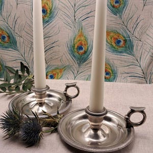 May include: Two silver candle holders with white candles. The candle holders have a round base with a handle and a decorative edge. The background is a fabric with a peacock feather pattern.