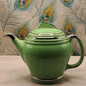 May include: A green ceramic teapot with silver detailing on the lid, spout, and base. The teapot has a curved handle and a lid with a fin-shaped design. The background features a fabric with a peacock feather pattern.