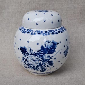 May include: A white ceramic ginger jar with a removable lid, featuring a hand-painted blue floral pattern of large chrysanthemum blossoms and leaves, accented by a delicate border of small blue dots scattered across the rounded body and lid.