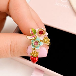 Può includere: Un anello color oro con un gruppo di fiori smaltati bianchi, azzurri e verdi, accentuati da piccoli cristalli rossi e rosa. Un piccolo ciondolo a forma di fragola smaltata rossa con semi color oro pende dalla composizione floreale.