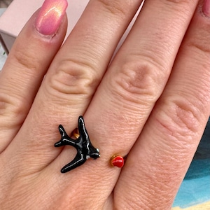 Puede incluir: Un anillo abierto de tono dorado que presenta un motivo de golondrina negra brillante en un extremo y una pequeña forma de corazón rojo liso en el otro. El anillo está diseñado para rodear el dedo con el pájaro y el corazón frente a frente.