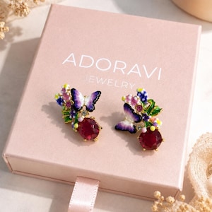 Puede incluir: Un par de pendientes dorados con mariposas de esmalte morado y blanco, pequeños detalles florales de colores y una piedra roja redonda facetada. Los pendientes se presentan sobre una caja rosa claro con el texto ADORAVI JEWELRY en blanco.