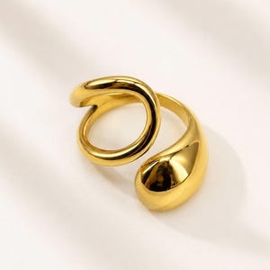 May include: A polished gold-tone ring featuring an open, bypass-style design. The band consists of a smooth, rounded wire that loops into a circular shape on one side and terminates in a larger, teardrop-shaped solid element on the other side.