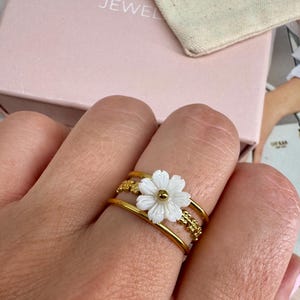 May include: A gold-colored ring featuring a white flower with a gold center. The ring has a triple-band design with decorative gold accents. The ring is on a finger, with a pink jewelry box in the background.