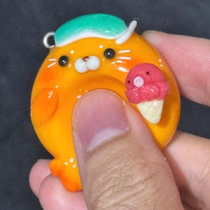 May include: A round, orange, glossy charm featuring a stylized character with a green sleeping mask on its head and a small pink ice cream cone held in its hand. The character has black eyes, a white muzzle, and white dots on its lower body.