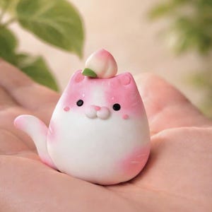 Peut inclure: Une figurine de chat rose et blanc fantaisiste, ornée d'une pêche. Le chat a des yeux noirs, des joues roses et une queue enroulée. La pêche a une feuille verte. La figurine est tenue dans la main d'une personne.