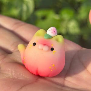 Könnte beinhalten: Eine kleine, abgerundete Katzenfigur mit einem Farbverlauf von Gelb an den Ohren bis zu einem leuchtenden Pink an der Basis. Sie hat zwei schwarze Augen, eine kleine rosa Nase und eine winzige weiße Erdbeere mit einem grünen Blatt auf dem Kopf.